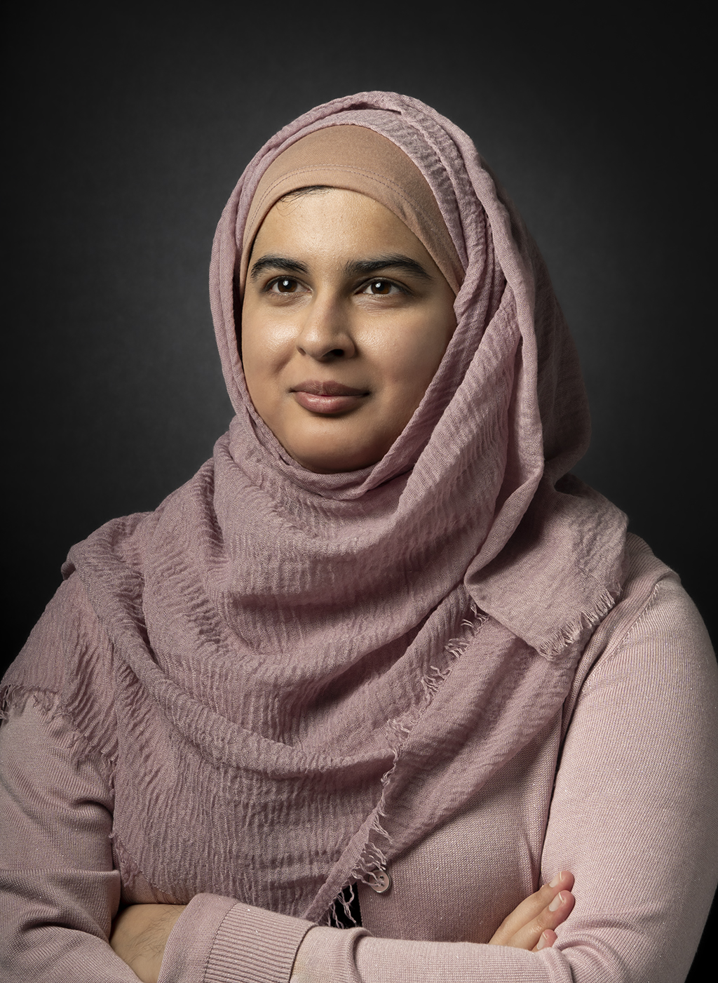 Business portrait of a person in a pink hijab and cardigan on a black background looking off-camera to the top left and smiling.
